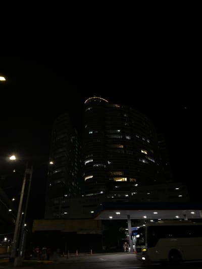 This nighttime image depicts an urban scene in Panama City, Panama. Two high-rise buildings dominate the background against a dark sky. The taller building on the right features a curved or wavy facade with numerous illuminated windows and a brightly lit crown structure at its apex. The name "BAY VIEW" is visible on a lower, wider section of this building. A second, darker high-rise structure is partially visible to the left. In the foreground, a brightly illuminated gas station canopy with multiple overhead lights is positioned on the right side of a multi-lane asphalt road. A white bus with illuminated headlights is partially visible beneath or adjacent to the canopy, appearing to be in motion or recently stopped. On the left, multiple streetlights emit bright, yellowish-white light from poles lining the road. A group of approximately five individuals is visible on the sidewalk in the lower-left quadrant, near dark barriers or fencing. Other indistinct figures are present near the gas station area. The overall environment is dark, with artificial light sources providing illumination.