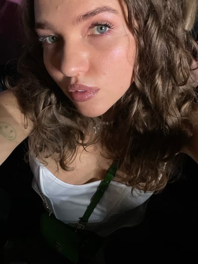 A selfie of a woman with light skin, blue eyes, and curly brown hair. She is wearing a white top and a green purse with a matching strap. A small cross earring is visible. The image is taken at an angle with a dark background. She has a small smiley face tattoo on her arm. Her lips are glossy.