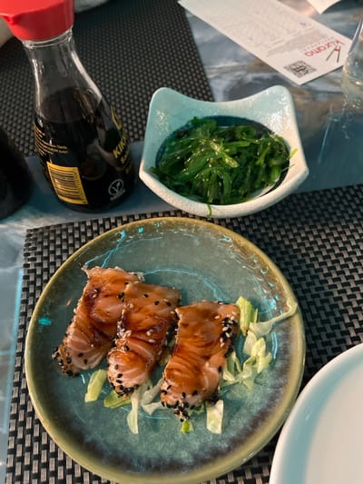 An overhead shot captures a dining scene at what appears to be a restaurant in Basiglio, Italy. The table is set with a dark, textured placemat.

The primary subjects are food items. On the left, a small, white, uniquely shaped bowl holds a vibrant green seaweed salad, sprinkled with sesame seeds. To its right, a round, light blue-green ceramic plate features three pieces of what looks like seared or glazed salmon, topped generously with black and white sesame seeds, and garnished with shredded lettuce. Part of an empty white plate is visible in the upper right background.

Other objects on the table include a clear drinking glass, mostly obscured, and a Kikkoman soy sauce bottle lying on its side in the lower-left foreground. A paper menu or receipt, featuring the name "Kurama" and a QR code, is also visible near the top-left corner.

The setting suggests a meal is about to be enjoyed, likely at an Asian-fusion or Japanese restaurant, given the food items and soy sauce. The lighting is ambient, indicating an indoor environment, but the exact time of day is not discernible. No people are visible, and no specific emotions are conveyed. The visible text includes "Kurama" on the menu and "Kikkoman" and "Soy Sauce" (or "Salsa di Soia") on the bottle, along with a barcode and other small print.