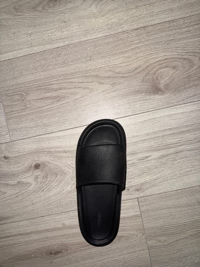 A single black slide sandal lies flat on a light-colored wooden floor. The sandal is positioned vertically in the lower center of the frame, with its toe pointing upwards. The wood grain of the floor is visible, with planks running horizontally. The scene is devoid of any human presence or other objects. The location is Hainstadt, Germany.