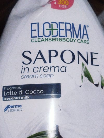 An upward, close-up shot shows the label of a white plastic bottle. At the top, a pink oval with a white drip and yellow text reads "300 DOSI". Below this, the brand name "ELC DERMA" is printed in large blue letters with a green cross in the "C". Underneath, "CLEANSER & BODY CARE" is written in smaller green text. The largest text on the bottle reads "SAPONE" in bold navy blue font, followed by "in crema" in smaller navy blue italics, and then "cream soap" in even smaller navy blue italics. To the left, a dark blue rectangular label contains white text that says "Fragranza Latte di Cocco" on the top line and "coconut milk" on the bottom line. Below this, a blue leaf logo is beside the words "Dermo Testato" in blue text. The bottle's surface has a slightly grainy texture.