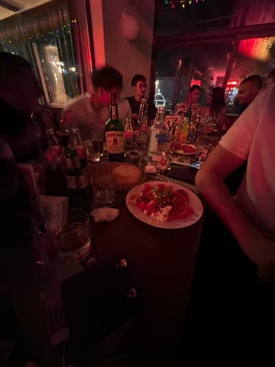 The image depicts a dimly lit bar in Panagyurski kolonii, Bulgaria, with a group of people socializing. The focus is on a table covered with numerous glasses, bottles (including Jameson), and a plate of salad. Several people are visible around the table, some looking towards a window. The bar setting suggests a casual, social gathering in an urban environment.