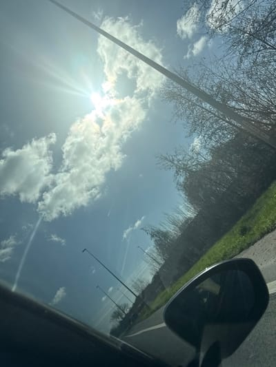 The image captures an outdoor scene on a bright, sunny day, likely viewed from the interior of a vehicle moving along a road in Lokeren, Belgium. The sky, predominantly blue, features scattered white cumulus clouds. The sun is prominently visible in the upper left quadrant, partially obscured by clouds, emitting distinct sunbeams. Multiple linear contrails from aircraft are present across the sky.
To the right, a dense line of bare deciduous trees and brush borders a patch of green grass, indicative of late winter or early spring. A dark, tall pole, likely a streetlight or utility pole, extends diagonally from the middle-right to the upper center, traversing the sky. Below, a paved road with a solid white edge line is visible. Several dark, slender streetlights are evenly spaced along the roadside, extending into the distant perspective.
In the lower right foreground, a portion of a dark car's interior, including the top edge of a dashboard or door panel, and the black casing of a passenger-side rearview mirror are visible. The mirror's reflective surface shows a blurred, dark reflection. No individuals are depicted.