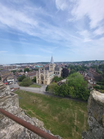 The image shows a wide-angle view of Rochester Cathedral and the surrounding cityscape of Rochester, United Kingdom. The cathedral, a prominent stone building with a tall spire, is centrally located. In the foreground, a grassy area is visible with a few people walking. Beyond the cathedral, the city extends with a mix of buildings, trees, and a glimpse of distant industrial structures and water. The sky above is blue with scattered white clouds. The perspective is from a high vantage point, possibly a castle wall, as indicated by the stone texture and rusty metal railing in the lower left and right corners of the image.