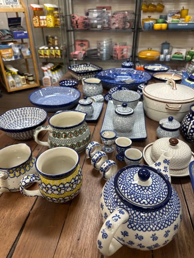This image depicts a display of Polish pottery, likely in a shop, with a focus on ceramic tableware. The pottery features intricate blue and white floral patterns, as well as some pieces with yellow floral accents. Visible items include teapots, mugs, bowls, serving dishes, a butter dish, and egg cups. The pottery is arranged on a wooden table, with shelves of other merchandise visible in the background, suggesting a retail setting. There are no people present in the image. The scene is indoors, with wooden flooring and shelving units. The lighting suggests ambient indoor lighting. The overall impression is one of a curated collection of decorative and functional ceramic items. The specific location in Bellingen, Australia, is not deducible from the image itself.