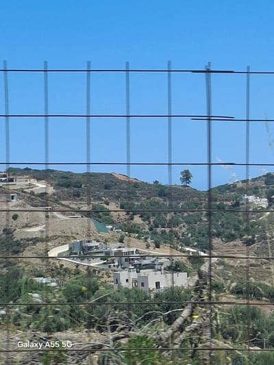 The image shows a landscape view through a window or other gridded structure. It includes a mountain with buildings and vegetation on its slopes. The sky is visible on the left, and the vegetation appears denser on the right side of the frame. The scene is identified as Vlichada, Greece, based on the camera's branding.