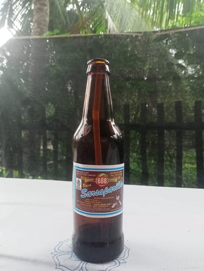The image depicts a brown glass bottle of "Sarsaparilla" beverage, positioned centrally on a white, textured surface, likely a tablecloth, adorned with a light blue patterned detail in the foreground. The bottle contains a dark liquid and features a full-color label.
The label prominently displays "Rasa Sarsaparilla" (Sarsaparilla Flavor) and the brand "Merek 688" with an apple-like graphic. Essential information on the label includes a production permit number "DIN. KES. P-IRT No. 113117413 0022-25," a "HALAL INDONESIA" logo, net content of "Isi Bersih 280 ml," a registration number "Reg. IDM 000109327," and an expiration date of "EXP. DECEMBER 2025." The product is identified as "MINUMAN BERKALORI RENDAH" (Low-calorie drink) and "DIPRODUKSI OLEH: PERUSAHAAN LEMOAN " ASIA MON 688 "" with the manufacturing location specified as "LHOKSEUMAWE - NAD INDONESIA" along with a detailed street address. A harp-like symbol is embossed on the glass bottle above the label. A transparent rod-like object is visible vertically inside the bottle, extending from the neck almost to the base.
The background, rendered in a shallow depth of field, depicts an outdoor environment, consistent with the location in the city of Teupinjok, Indonesia. Immediately behind the bottle, a dark, possibly mesh or woven, fence runs horizontally. Beyond the fence, out-of-focus green foliage and tree elements are visible, suggesting a natural or cultivated garden setting. A distinct tree trunk, possibly a palm tree, is visible on the far left. The scene is illuminated by diffuse natural light, creating bokeh effects in the background. No individuals are present in the image.