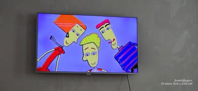 The image is a photograph of a television screen displaying a cartoon with three characters. The screen is mounted on a gray wall. The cartoon characters have yellow faces with large eyes, and distinct hairstyles and clothing. The character on the left has bright orange hair and is wearing a red shirt with a striped scarf. The character in the middle has yellow-green hair and a red and white striped shirt. The character on the right has pink hair and is wearing a blue and purple striped shirt. There is text on the screen in Russian, and a 0+ age rating. In the bottom right corner of the image, there is a watermark with text in English and Russian that reads "frenki4fingers 29 июня 2026 г. 8:06 AM". The location is identified as Verkhniy Naur, Russia.