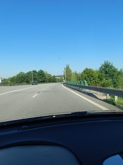 The image shows a road with a guardrail on the left side, lined with trees. A blue car is visible driving on the road. The sky is clear and blue, indicating a sunny day. The scene appears to be a highway or a similar type of road, with streetlights present. The perspective suggests the photo was taken from inside a vehicle. The image is oriented vertically. There is no visible text in the image. Based on the location context, the scene is likely in Písek, Czechia. The time of day appears to be daytime, given the bright sunlight.