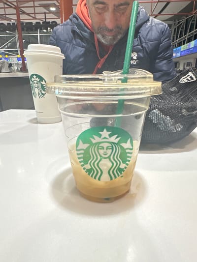 A man in a dark blue jacket and black baseball cap sits at a table with two Starbucks drinks. One is a tall, clear plastic cup with a green straw, lid, and the Starbucks logo visible. The other is a smaller white cup, also with the Starbucks logo. The scene appears to be indoors. The location is Pudahuel, Chile.
