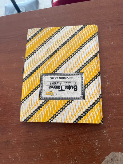 A top-down medium shot shows a notebook with a yellow and white batik pattern on a wooden table. The notebook is positioned in the center of the frame, at a slight angle. The batik pattern consists of diagonal rows of stylized floral motifs in yellow and white, separated by thin black lines and small diamonds. On the front cover of the notebook, there is a rectangular label with text. The label has a decorative border and contains text in Indonesian, with "Cap VISION BATIK" printed above a larger, handwritten word that appears to be "Buku Tamu," meaning guest book. Below "Buku Tamu," "VISION BATIK" is printed in a smaller font. The wooden table is brown and shows signs of wear, with some dust and lighter-colored scuff marks. In the upper left corner of the image, a portion of a white object with a black handle is visible, possibly a piece of furniture or an appliance. The overall lighting suggests an indoor environment. This image is of an object located on a table in Boja, Indonesia.