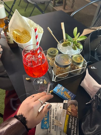 A table setting features an Aperol spritz with red and white striped straws, a cone of potato chips, a container with olives, and a three-tiered spice rack. A hand holding a lit cigarette sits near rolling papers and a cigarette pack with a warning label. A Marlboro cigarette pack, also with a warning label, is present. The setting includes beer bottles, a black handbag, and a silver ring.