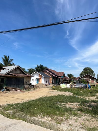 The image depicts a roadside scene in Dalam, Indonesia, under a bright blue sky with scattered wispy clouds, suggesting a sunny day, likely midday given the shadows. The viewpoint is from the side of a dirt road, looking towards a row of houses and vegetation.

The foreground is dominated by a dirt path bordered by patches of green grass and some concrete. Several power lines stretch across the sky on the left side of the frame, secured by poles.

The main subjects are a collection of houses, some appearing to be under construction or in a state of disrepair. The houses have various roofing materials, including corrugated metal and tiles, and some walls are painted in different colors, with one house featuring a white facade with orange and blue accents. A silver car is parked near one of the houses. Among the houses, there are also indications of building materials, such as piles of sand and wood. Palm trees are visible, a common feature in tropical locations like Indonesia.

The scene suggests a residential area, possibly a village or a less developed part of a town. There is no clear activity or event taking place; it appears to be a static view of the environment. The overall impression is one of a typical, everyday setting in a warm climate, with a focus on the built environment and the natural elements. No discernible text is visible in the image.