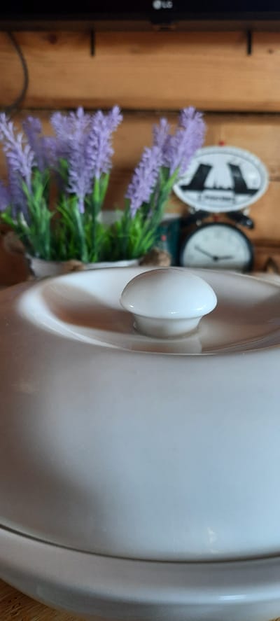 The image shows a close-up of a white lid with a round knob on top, part of a larger ceramic pot or dish. In the background, a blurred arrangement of artificial lavender flowers in a white pot is visible. Behind the flowers, a vintage-style mechanical scale with a white face and black hands is partially visible, and an oval decorative object with a silhouette of buildings is affixed to it. The backdrop appears to be made of wooden planks. The lighting suggests an indoor setting. The image does not provide any information about the city of Borogontsy, Russia, or any specific location.