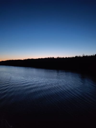 The image depicts a body of water, likely a lake or river, under a twilight sky. The sky transitions from a deep blue at the top to a faint orange glow on the horizon. Silhouetted trees line the distant shore. The water's surface shows subtle ripples and disturbances, suggesting a slight breeze or current. The scene is dimly lit overall. It is not possible to definitively locate the scene in the city of Uzhla, Russia, or ascertain any details about people or actions from the image alone.