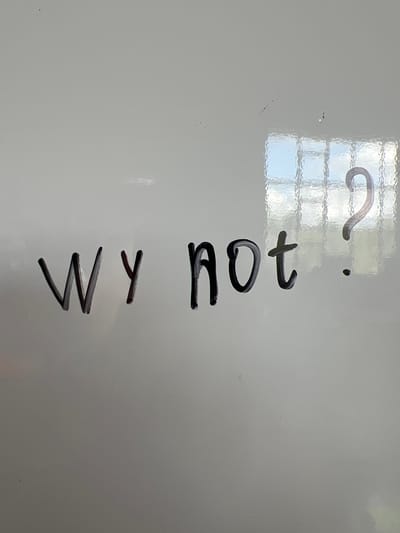 The image is a close-up, eye-level shot of a whiteboard with the words "wy not?" written in black marker. The lettering is somewhat smudged and appears to be written in a casual, handwritten style. To the right of the text, a question mark is drawn, followed by a small dot. A reflection of a window with many panes is visible on the surface of the whiteboard, indicating it's likely indoors. The overall background is a plain, white surface, consistent with a whiteboard. The image does not depict any people, animals, or discernible city landmarks. The text on the whiteboard is the sole subject of the image.
