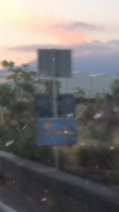 The image shows a blurry outdoor scene at dusk, with a soft orange and pink sky. In the background, several buildings with white roofs are visible. In the mid-ground, there is a large, dark, rectangular structure, possibly a building or storage unit. To the left of this structure, there are trees and foliage. In the foreground, a blue rectangular sign with yellow markings is partially visible. The overall impression is that the photo was taken from a moving vehicle, as indicated by the motion blur and the presence of what appear to be specks of dust or water droplets on the surface from which the photo was taken. The location is identified as Belpasso, Italy, though no specific landmarks or features definitively confirm this without further context.