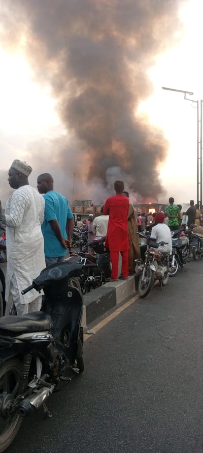 A group of people stand near a road, observing a fire and plumes of smoke. Parked motorcycles line the road, positioned on the curb. Some individuals are dressed in traditional attire, while others wear more casual clothing. The smoke rises high, obscuring the details of the building where the fire appears to be located. A streetlight pole is visible on the right edge. The overall scene indicates a serious event drawing the attention of onlookers.
