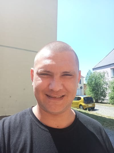 A bald man in a black t-shirt is pictured outdoors in Bitterfeld-Wolfen, Germany. The man is smiling and looking directly at the camera. He has fair skin and appears to be in his 30s or 40s. The background shows a building with a light-colored wall, a bright blue sky, and some trees and a yellow car parked on the side of a road. The sun is shining brightly, casting shadows on the ground. The overall impression is of a casual, sunny day.