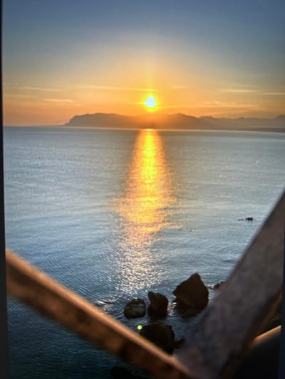 The image shows a sunset over the sea, with a coastline visible in the distance. The sun is low on the horizon, casting a bright reflection on the water. There are rocks in the foreground, and some blurred elements that suggest a railing or structure. The location is likely Castellammare del Golfo, Italy, given the characteristic shape of the coastline and the presence of the sea. No people are visible in the image. The overall scene is serene and captures the natural beauty of a coastal sunset.