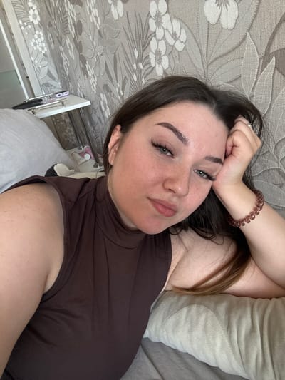 A young woman with dark hair and green eyes lies on her side, looking towards the camera. She is wearing a brown, sleeveless, high-necked top. Her right arm is bent, with her hand resting on her forehead, and she is wearing a ring on her index finger and a coiled bracelet on her wrist. She has dramatic false eyelashes and appears to be wearing makeup. Her skin has a scattering of freckles.
The woman is lying on a bed with a light-colored, textured blanket or duvet. Behind her is a headrest or pillow, also in a light color with a textured pattern. To her left, the background features a wallpaper with a floral pattern in gray and white tones. On the far left of the frame, a white shelf unit is visible, with a few items on it, including what appears to be a toothbrush in a holder and some small bottles or containers.
The lighting suggests it is daytime. The overall mood appears relaxed or casual. The location context provided is Гамово, Russia. There is no visible text in the image.