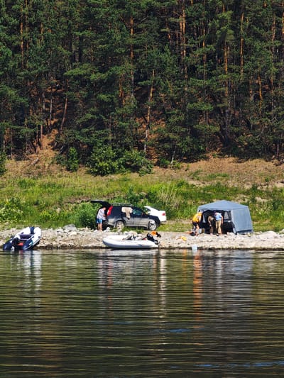 The image depicts a riverside scene in Podporozh'e, Russia, characterized by a dense coniferous forest bordering a rocky shoreline. In the foreground, the water's surface is rippled and reflects the surrounding environment. On the shore, there are several inflatable boats, with one partially submerged in the water. A blue and grey tent is set up, with several individuals gathered inside and around it, engaged in activities that suggest camping or preparing for water excursions. Nearby, a black SUV with its trunk open is parked, and its contents are partially visible. The overall impression is of a recreational outing in a natural setting, with elements of outdoor activity and logistics evident.
