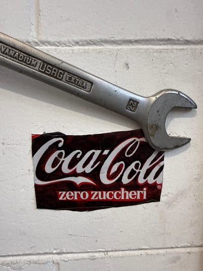 The image shows a close-up view of a metal wrench resting on a white brick wall. The wrench has "VANADIUM USAG EXTRA" and the number "27" etched into it. Below the wrench, a portion of a Coca-Cola Zero Zuccheri label is visible. The label is dark red and black with the Coca-Cola logo in white. The wall appears to be outdoors, possibly in an industrial or workshop setting. The lighting suggests it is daytime. The image is taken from a slightly elevated angle, looking down at the objects. No people are visible in the image. The location is identified as Fontevivo, Italy.