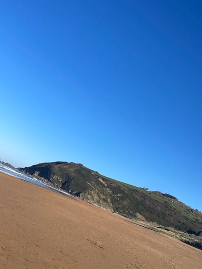 The image captures a sunny coastal scene in Zarautz, Spain, presented from a tilted perspective. Approximately two-thirds of the frame is occupied by a clear, deep blue sky, devoid of clouds.

The lower third depicts a wide, reddish-brown sandy beach that angles upwards from the lower left to the middle right. On the far left, white-capped waves are visible breaking on the shoreline where the sand meets the ocean.

In the mid-ground and background, a large, dark green, heavily vegetated hill or mountain (Monte San Antón) rises from the water on the left, forming a prominent natural landmark. Its slopes are covered with varied green foliage and some visible rocky outcrops. Further to the right along the base of this hill, a line of wooden posts or fencing runs parallel to the beach.

A single, small, dark-silhouetted individual is observed walking on the sand in the mid-ground, positioned near the base of the hill on the right side of the visible beach area. The entire scene is brightly illuminated by natural sunlight, indicating daytime conditions.