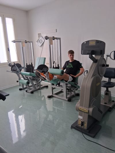 A young male is using a weight machine in a gymnasium. He is wearing a black t-shirt with a green logo and white shorts. The gym is brightly lit and has a green tiled floor. There are other weight machines visible in the background, as well as a window. The user is sitting on a padded seat and his legs are positioned under a padded bar. He appears to be in the process of performing a leg exercise. Visible text on the weight machine includes "TECHNOGYM" and "SILVER". The time of day is not discernible.