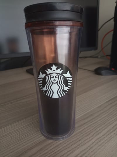 The media file shows a Starbucks tumbler placed on a wooden desk. The tumbler is made of clear plastic with a dark brown or black lid. Inside the tumbler, there appears to be a dark liquid, likely coffee or tea. A prominent Starbucks logo, featuring a siren, is visible on the side of the tumbler. The background suggests an indoor setting, possibly an office or home, with a computer monitor and a computer mouse visible. The lighting indicates it is daytime. The overall scene is neutral and focuses on the product. There is no visible text other than the Starbucks logo. The location context is Ankara, Turkey.