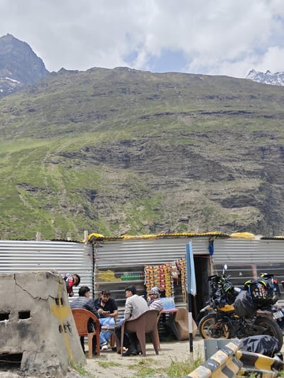 A group of four men are seated at tables outside a makeshift roadside shop in a mountainous region of India. The shop is constructed of corrugated metal with a yellow tarp roof. Shelves inside display snacks and drinks. A motorcycle, heavily loaded with luggage, is parked to the right of the shop. In the background, a steep, rocky mountain slope covered in sparse green vegetation rises under a cloudy sky. The man on the far left is wearing a grey jacket and a beanie. The man next to him, wearing a black and white striped jacket, is looking down at his food. The man in the center, wearing a black t-shirt and blue jeans, is also looking at his food. The man on the far right, wearing a grey jacket, is looking to his left. Another man in a black and red jacket is standing and bending over a table to the left of the seated men. A large, cracked concrete structure with square openings is in the foreground on the left.