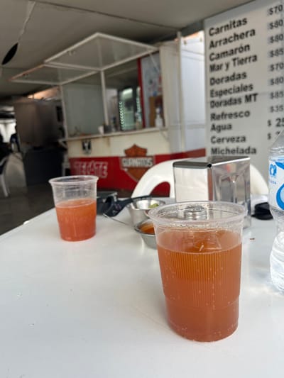Two translucent plastic cups filled with an amber liquid are situated on a white table. A menu board displays prices for various food items, including Carnitas, Arrachera, and Camarón. A metal napkin holder and a bottle of water are also visible. Behind the table, a food stall with a red sign that reads "TEGATE GUANATOS" is apparent. The stall has a white framework, and interior shelves are visible.