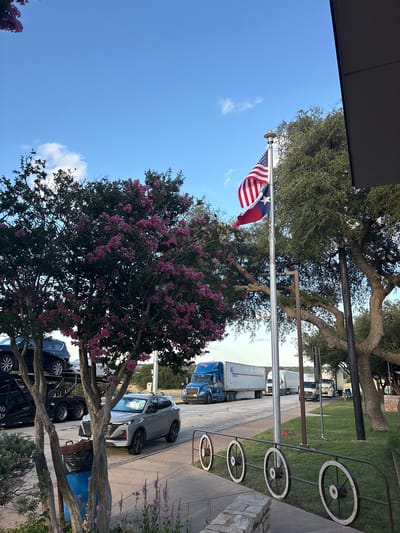 The image depicts a scene at a rest stop on I-10 in Sonora, Texas, United States. The foreground features a sidewalk with decorative wagon wheel railings and a stone border. A light gray SUV is parked on the asphalt. To the left, a car carrier truck is partially visible, loaded with dark-colored vehicles. A trash bin with a blue liner is situated next to a tree with purple flowers.
In the mid-ground, several large semi-trailer trucks are parked on the side of the road, with a blue truck prominently displayed. A flagpole in the center of the frame flies the American and Texas flags. A mature oak tree with large, gnarled branches is visible behind the flagpole. The background shows a clear blue sky with scattered white clouds, and more trees and distant vegetation. The lighting suggests it is either early morning or late afternoon.