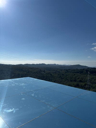 A landscape featuring a clear blue sky with a few clouds, transitioning to a distant horizon line of mountains. Lush green forest covers the terrain. Power lines stretch across the scene. To the right, a building or structure composed of mirrored panels reflects the sky and surrounding environment. The image is oriented sideways, presenting a unique perspective of the natural and built environment.