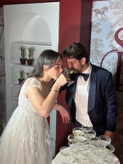 A newly married couple is toasting with champagne. The bride is in a white gown, and the groom is in a suit. They are standing next to a tower of champagne glasses. The location appears to be a reception hall in Orizaba, Mexico, with a maroon wall above a white wall with small green plants. The lower wall has wallpaper with a brown pastoral pattern.