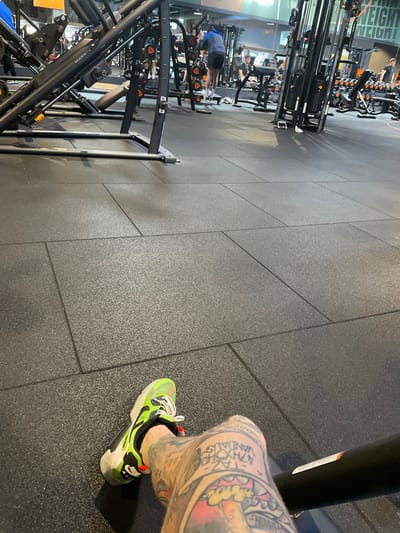 The image depicts the interior of a Basic-Fit gym in 's-Hertogenbosch, Netherlands, observed from a low, first-person perspective. In the immediate foreground, the right leg of an individual is visible from the mid-thigh downwards, resting horizontally. The leg is heavily tattooed with various designs, including a stylized mushroom and text reading "TAZ 4PSYCHO VANDALISM". A vibrant neon green, black, and white athletic shoe with red laces is worn on the foot. The leg rests on a black horizontal bar, likely part of a gym machine. The floor consists of dark grey, textured interlocking rubber mats.
The background shows a spacious gym environment containing numerous pieces of fitness equipment. Prominent machinery includes black and grey weight machines, such as a leg press machine marked "MATRIX" on the left, and various cable machines and dumbbell racks on the right. Several individuals are present, engaging in physical activity. One male subject in a blue hoodie and black shorts is observed near a weight rack in the mid-left background. Other figures are indistinctly visible further back. The gym is well-lit by overhead artificial lighting, and a wall in the background features the partial branding "BASIC-FIT" and "WEIGHT".