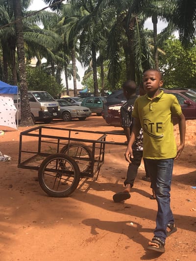 Two young boys stand on a red dirt ground in Nteje, Nigeria. One wears a yellow shirt and jeans, the other a black and white patterned shirt with black pants. They stand next to a metal-framed cart with bicycle wheels. Behind them, a row of cars and vans are parked, and tall palm trees line the scene's left side.