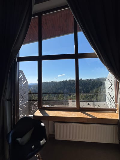 View from a window with brown wooden frame in a room, showing an outdoor scene with a lush, green forest, blue sky, and parts of a modern concrete building with a design of cut-out squares. A partially drawn curtain is visible on the left side of the window. In the room, a white radiator is attached to the wall next to the window, and part of a black chair is visible at the bottom right. The scene is likely in Pamporovo, Bulgaria, based on the context.