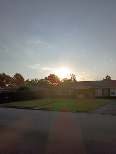 The image depicts a suburban street scene at sunset in Richmond, United States of America. The sun is low in the sky, casting a warm glow and creating lens flare effects across the foreground. In the middle ground, a row of single-story brick houses with green lawns and mature trees in front of them is visible. The sky is a pale blue with scattered wispy clouds. The street in the foreground is paved and appears empty.
