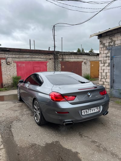 A grey BMW 650i coupe is parked on a dirt and gravel surface in front of a row of brick garages in Veteran-2, Russia. The car is facing away from the camera, with its rear visible. The license plate reads "X677YY 18 RUS". The garages have various colored doors, including red, brown, and yellow. The sky is overcast and cloudy. The environment appears to be an outdoor storage or parking area.