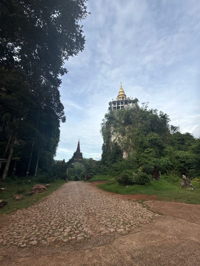 A cobbled stone pathway leads towards an ancient arched gateway covered in dense green foliage. Beyond the gateway, a steep, lush green cliff rises dramatically, crowned by a multi-tiered golden stupa. The sky above is partly cloudy with a muted, overcast light. The scene is set within the natural landscape of Ban Sadet, Thailand, with tall, dark green trees flanking the left side of the pathway. The overall impression is one of serene, natural beauty with a spiritual monument at its peak.
