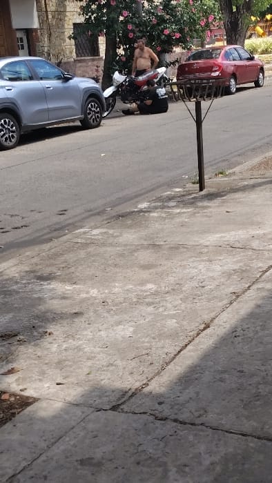 A man stands shirtless next to a red and white motorcycle, parked on a street beside a silver car. Lush greenery with pink flowers frames the scene on the left. Two cars are visible further up the road. The street is concrete with visible cracks and uneven textures. A shadow covers a large portion of the right side of the frame.