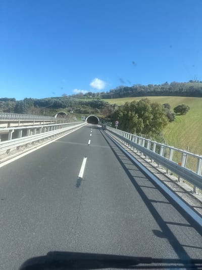 The image depicts a multi-lane asphalt highway on a clear, sunny day, leading towards two tunnel entrances within a hilly, vegetated landscape near Martinsicuro, Italy.

The roadway, observed from a forward-moving perspective, exhibits two clearly marked lanes separated by white dashed lines, with a solid white line bordering the right lane. Metal guardrails line both sides of the visible highway segment. On the left, a parallel, elevated concrete bridge structure is partially visible. The road proceeds directly into a dark, circular tunnel entrance. To the left of this primary tunnel, a second, wider tunnel entrance, partially obscured by vegetation, is also visible.

The surrounding environment consists of undulating green hills covered with sparse deciduous and evergreen trees, as well as cultivated fields. Shadows from the right guardrail are cast diagonally across the road surface, extending towards the left. The sky is a vivid, cloudless blue with a single small white cumulus cloud visible on the right horizon. No people or active vehicles are present within the frame, aside from the implied vehicle from which the photograph was taken, evidenced by a dark shadow at the bottom of the image. Minor lens flares or dust particles are visible in the upper sky area.