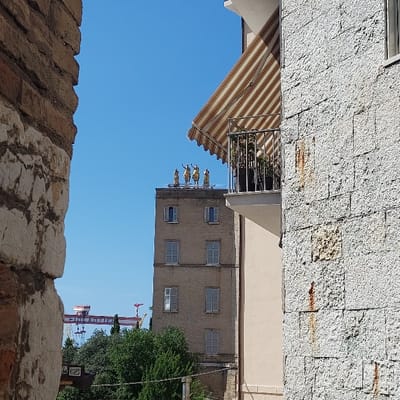 The image depicts a detailed daytime urban scene in Ancona, Italy, viewed through a narrow vertical opening framed by two structures.

On the left, a wall constructed from reddish-brown bricks and light-colored stone blocks occupies the immediate foreground. On the right, a light-colored, rough-cut stone wall is visible, displaying vertical rust stains. This wall features a balcony with a striped awning (beige and off-white) extended over its railing, holding several dark potted plants.

Centrally, in the mid-ground, stands a tall, multi-story building with a light beige facade, featuring multiple rectangular windows. Some windows are fitted with light-colored louvered shutters, appearing either partially open or closed. Perched prominently on the building's flat rooftop are four large, golden-toned equestrian statues, identified as the Quadriga dell'Aurora, characteristic of the Palazzo del Popolo in Ancona.

In the lower left background, green tree canopies are discernible. Further in the distant background, against a clear blue sky, industrial port structures are visible, including a prominent red gantry crane bearing the inscription "FINCANTIERI" and other vertical crane elements. The entire scene is brightly illuminated by natural daylight.