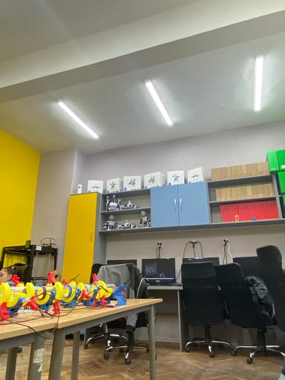 This image captures a vibrant and organized robotics or engineering workshop, likely a classroom or lab, in Vetrino, Bulgaria. The room features light grey walls, with a striking bright yellow accent wall visible at the lower right. The ceiling is equipped with multiple linear fluorescent light fixtures, providing bright illumination. The floor is made of wooden parquet.
Along one wall, a comprehensive shelving unit displays various items. The top shelves hold neatly stacked green boxes, possibly containing electronics or educational kits. Below these are shelves with cardboard boxes and a section with red binders or containers. Light blue cabinets with silver handles are positioned beneath these shelves. Further down, open shelving units showcase numerous white boxes, each depicting a robot, along with several assembled small robots, indicating a focus on robotics education or development. A yellow cabinet with a single door is at the bottom of this unit.
To the right, several black office chairs with wheels are arranged around workstations. Some chairs have dark jackets or bags draped over them. Two computer monitors are visible on desks, one of which has "KTC" printed on its bezel and displays a detailed 3D model, possibly a CAD rendering of a mechanical or robotic component. Multiple cables run from the computers to wall-mounted power outlets.
In the foreground, on a long wooden table, an elaborate robotics project is set up. It consists of interconnected yellow and blue plastic components, secured with red clamps and wired together, resembling a modular mechanism or a learning kit. A 3D printer with a black frame is partially visible on the table near the yellow wall. A person's head and shoulder are partially visible in the bottom right corner, actively engaged with the robotics project.
The scene suggests an environment dedicated to hands-on learning and practical application in technology or engineering.