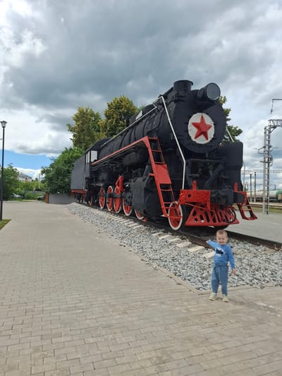 In Yanaul, Russia, a young child in a blue long-sleeved shirt and pants stands on a paved walkway, pointing towards a large, black steam locomotive displayed on railway tracks. The locomotive features prominent red accents on its wheels, ladders, and a large red star emblem on its front. The scene is set against a backdrop of green trees and a cloudy sky. The walkway is made of grey interlocking pavers, and the train is surrounded by gravel. In the background, other train cars and overhead power lines are visible, suggesting the location is near an active or historical railway area.