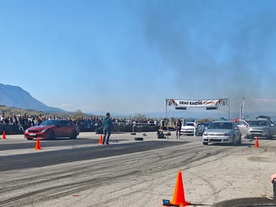 The image depicts a drag racing event taking place in Kraynitsi, Bulgaria. A red Mercedes is positioned on the left lane, facing the finish line. A grey Volkswagen is placed on the right lane. People are standing along the sidelines, observing the race. Orange traffic cones are positioned along the racetrack. A banner reading "DRAG RACING КРАНИМИЦИ" is visible over the finish line. Smoke is visible in the air. The road surface displays many tire marks. Mountains are visible in the background under a blue sky.