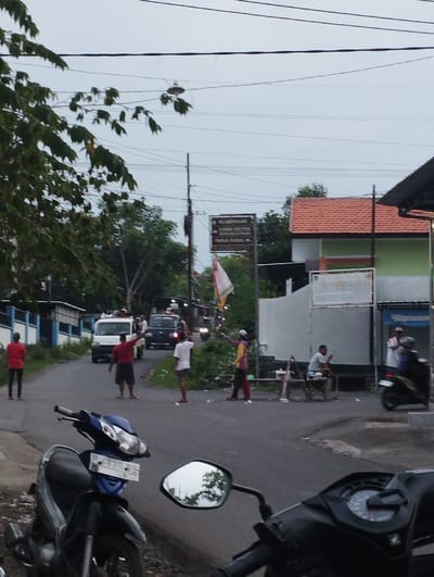 This media file captures an outdoor street scene, likely in a residential or semi-rural area in Indonesia. The image is taken from behind two parked motorcycles in the foreground, with the street curving to the left.
Numerous people are visible on and around the asphalt road. In the midground, several men are standing on the road, some appearing to direct traffic or signal with their hands. One man in a red shirt stands on the left side of the road. Another man in a white shirt and dark shorts points towards the distance, while a man in a striped shirt and dark pants on the right also signals. A white van and a dark car are further down the road, with other vehicles and people in the distance. Two men are seated on a wooden bench to the right of the road.
Notable objects include a street sign positioned on the right side of the road, a red and white flag (likely the Indonesian flag) attached to a pole near the sign, and various residential buildings with red tiled roofs and light-colored walls in the background. Overhead power lines crisscross the cloudy sky. Trees and foliage are visible on both sides of the street.
The sky is overcast, suggesting either early morning, late afternoon, or a heavily clouded day, contributing to the soft, even lighting across the scene. The activity appears to be a community gathering or event, possibly involving traffic management or a procession, given the people on the road and their coordinated gestures.
Visible text on the street sign includes directional information, with lines such as "NLAMPOK," "DESA SALAMAN," "DUKUH SENTONOSAN," and "PANJI KIDUL," providing specific location cues within the Kecamatan Panji area.