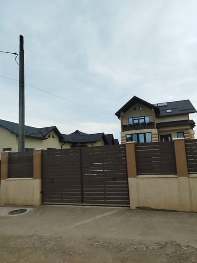 The image depicts a residential property in Băleni-Români, Romania, under an overcast sky. A tall, weathered utility pole stands to the left of the frame. In the foreground, a substantial, modern gate made of dark brown horizontal slats is prominently featured. This gate is supported by two beige concrete pillars. The gate opens onto a paved area that leads to a large, multi-story house. The house has a light-colored facade and a dark tiled roof with several prominent gables. Another smaller house with a similar roofline is partially visible to the left. The property is enclosed by a fence that complements the gate's design, consisting of dark brown horizontal slats atop a beige concrete base. The ground in front of the gate is a mix of gravel and concrete, with a manhole cover visible on the left. The overall impression is that of a well-maintained, contemporary suburban dwelling.