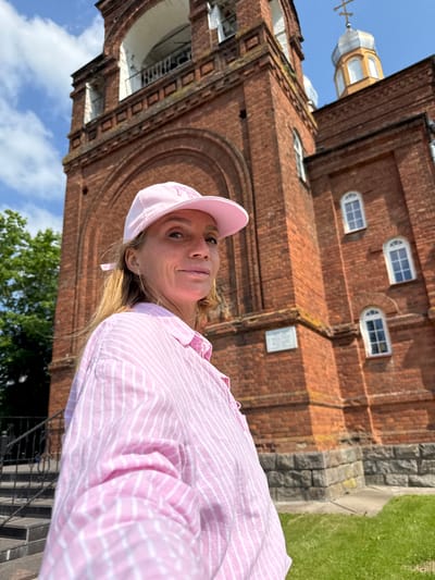 In an outdoor, low-angle, medium shot, a woman wearing a pink baseball cap and a pink and white striped button-down shirt is positioned in the foreground, facing right and slightly towards the camera. She has blonde hair and is looking at the camera with a slight smile. Behind her, a large, red brick building, identified as the Church of the Nativity of the Theotokos in Chudovo, Russia, dominates the background. The church features an arched entranceway, arched windows with white trim, and a silver dome topped with a cross. The sky is blue with scattered white clouds. To the left of the church, green trees are visible, and a set of stone steps with a black metal railing leads up to the building. The ground in front of the church is covered in green grass and stone.